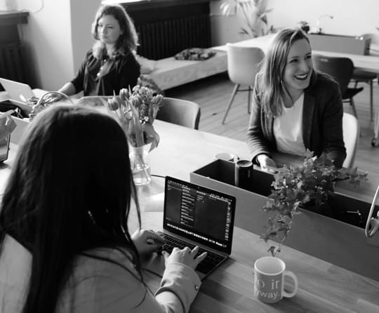 3 women sitting at a desk, in a modern office space, working on their laptop. One of them is smiling and has her mobile phone in her hands.