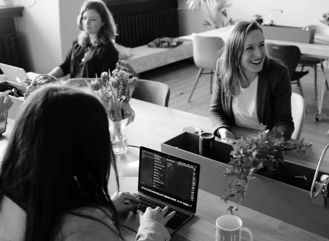 3 women sitting at a desk, in a modern office space, working on their laptop. One of them is smiling and has her mobile phone in her hands.